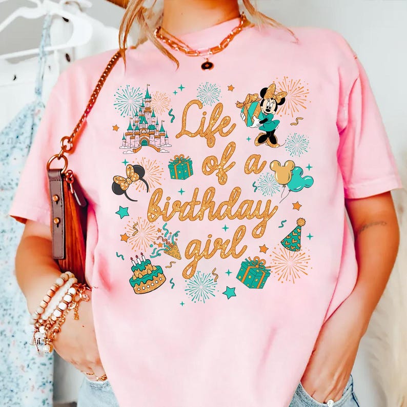 Pu&ograve; includere: T-shirt rosa chiaro con un design grafico colorato. Il design include un castello, Minnie Mouse, regali, fuochi d'artificio e il testo "Life of a birthday girl" in lettere dorate. Stile casual e confortevole.