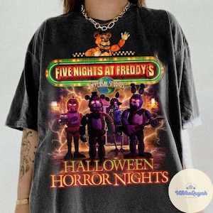 May include: Dark gray t-shirt featuring a graphic from "Five Nights at Freddy's" with the text "Halloween Horror Nights." The design includes several animated characters and the Universal Studios logo.