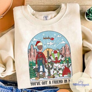 May include: Cream-colored sweatshirt with a graphic featuring two cartoon characters in a desert scene with a Christmas theme. The text "YOU'VE GOT A FRIEND IN ME" is printed at the bottom of the graphic.