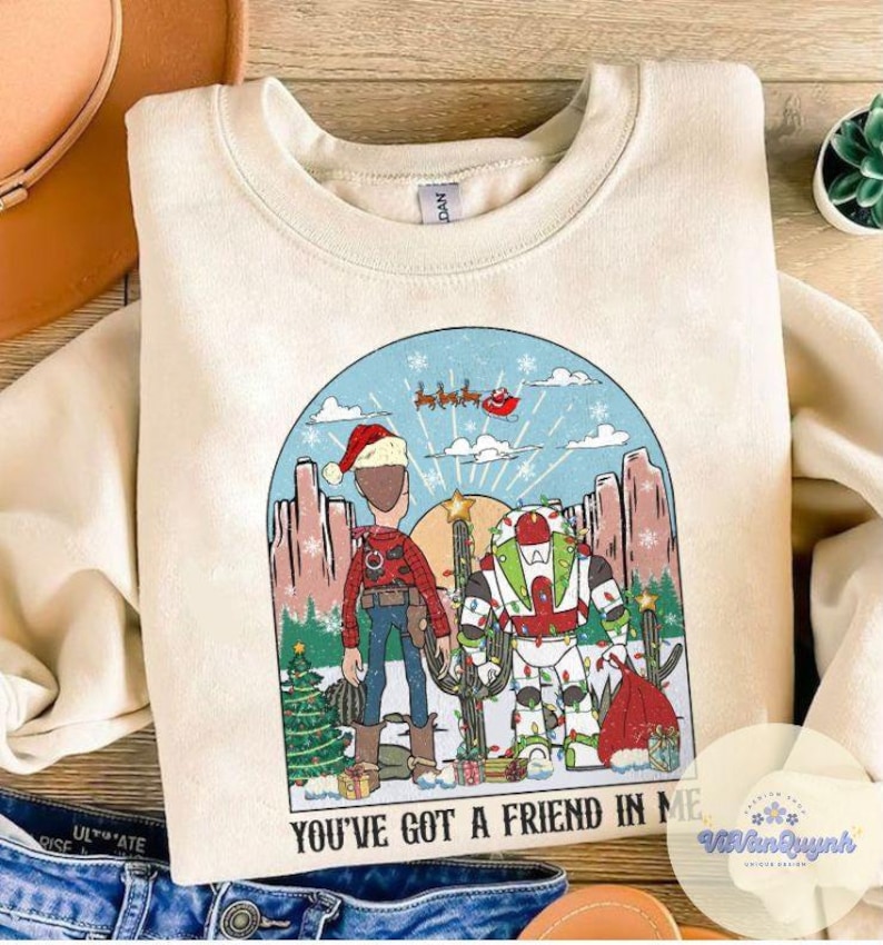 May include: Cream-colored sweatshirt with a graphic featuring two cartoon characters in a desert scene with a Christmas theme. The text "YOU'VE GOT A FRIEND IN ME" is printed at the bottom of the graphic.