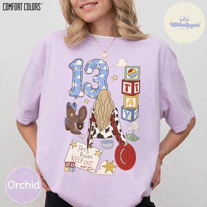 May include: A light purple short-sleeve t-shirt featuring a graphic print of the number 13, a person with long blonde hair, cowboy boots, toy blocks, and a sign that reads Tays Room KEEP OUT except you. The design includes a red hat and green alien.