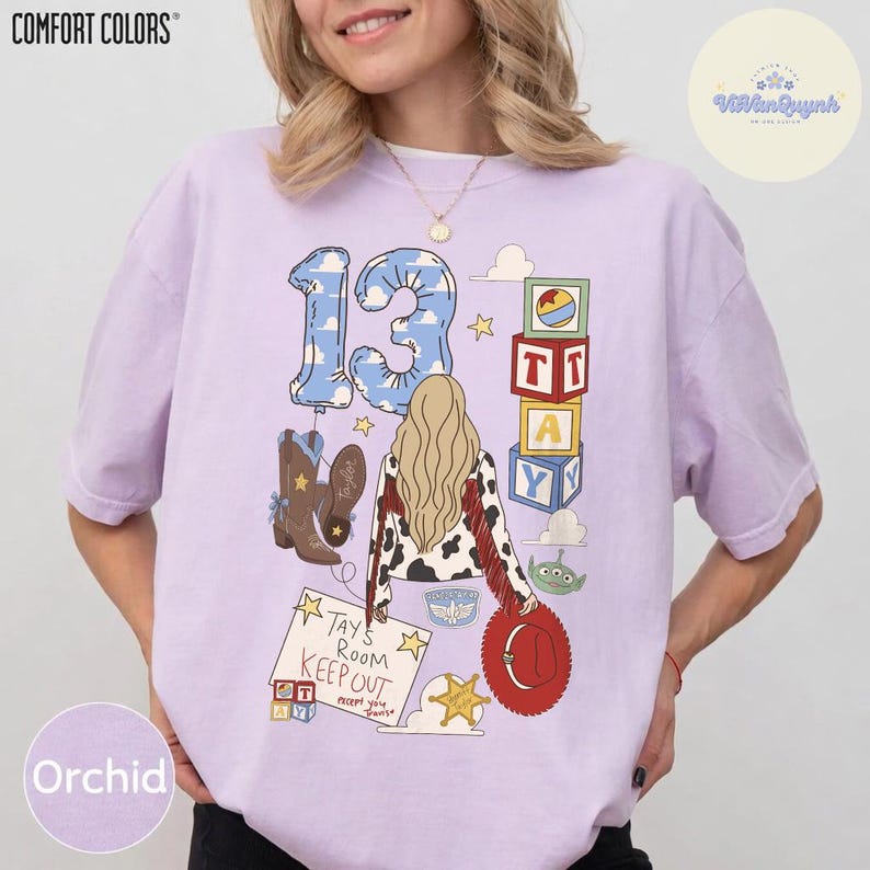 May include: A light purple short-sleeve t-shirt featuring a graphic print of the number 13, a person with long blonde hair, cowboy boots, toy blocks, and a sign that reads Tays Room KEEP OUT except you. The design includes a red hat and green alien.