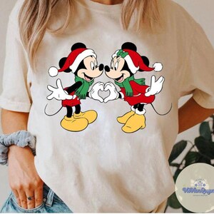 Op de afbeelding: Cr&egrave;mekleurig T-shirt met een afbeelding van Mickey en Minnie Mouse in kerstkleding. Ze dragen kerstmutsen, rode jasjes en groene sjaals en vormen met hun handen een hart. Het ontwerp is gecentreerd op het shirt.