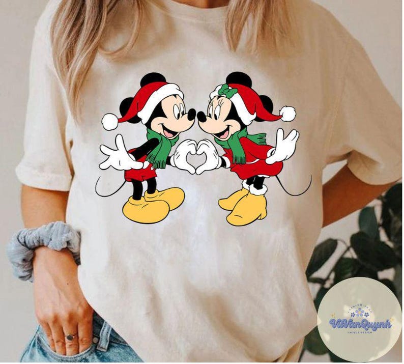 Op de afbeelding: Cr&egrave;mekleurig T-shirt met een afbeelding van Mickey en Minnie Mouse in kerstkleding. Ze dragen kerstmutsen, rode jasjes en groene sjaals en vormen met hun handen een hart. Het ontwerp is gecentreerd op het shirt.