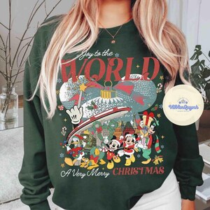May include: Dark green sweatshirt with a festive design featuring Mickey Mouse, Donald Duck, and Goofy. The graphic includes the text "Joy to the WORLD" and "A Very Merry CHRISTMAS" in red and white. A holiday-themed sweatshirt.