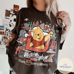 May include: Dark gray t-shirt featuring a graphic of Winnie the Pooh in a Santa hat holding a candy cane, with the text "Walt Disney World." The design includes various Disney World landmarks and snowflakes.