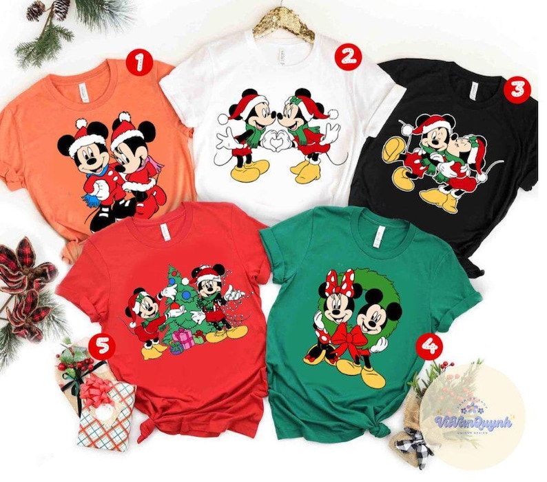 Op de afbeelding: Een verzameling van vijf t-shirts met korte mouwen met Mickey en Minnie Mouse in kerstthema's. De shirts zijn in verschillende kleuren, waaronder oranje, wit, zwart, rood en groen. Elk shirt heeft een andere afbeelding van de iconische personages.