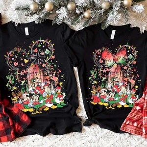 May include: Two black t-shirts with a colorful graphic featuring Mickey Mouse and friends in a Christmas theme. Red and black plaid pajama pants and red pajama pants with a white bear pattern are also visible.