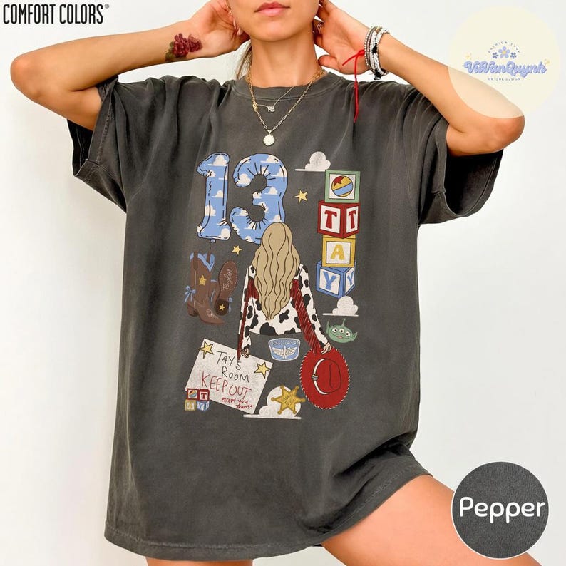 May include: A charcoal gray short-sleeve t-shirt featuring a graphic print with the number 13, a person with blonde hair, alphabet blocks spelling TAY, a brown cowboy boot, a red hat, and a sign that reads TAY'S ROOM KEEP OUT except you.