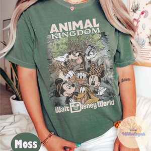 K&ouml;nnte beinhalten: Moosgr&uuml;nes T-Shirt mit einer Grafik von Disney-Figuren, darunter Mickey, Minnie, Donald und Goofy, mit dem Text "ANIMAL KINGDOM" und "Walt Disney World". Das Shirt hat einen Vintage-Look und ist leicht verwaschen.