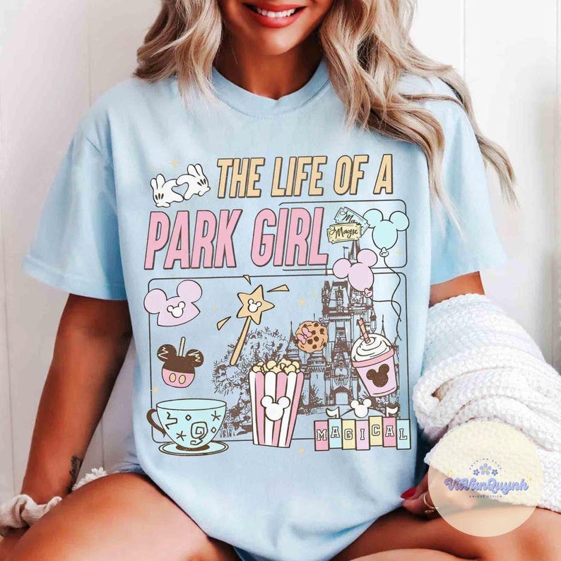 May include: Light blue t-shirt with the text "THE LIFE OF A PARK GIRL" and various cartoon graphics. The graphics include a castle, popcorn, a teacup, and Mickey Mouse-themed designs. The shirt has short sleeves.