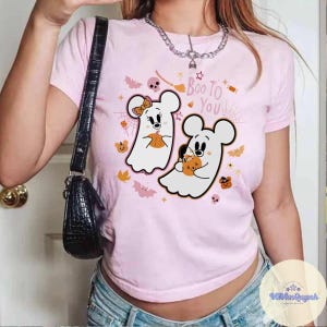 May include: A light pink t-shirt featuring a Halloween-themed graphic. The graphic includes two ghost-like figures resembling cartoon mice, each holding a pumpkin. The phrase "Boo To You" is written above the figures, with bats and spiderwebs.