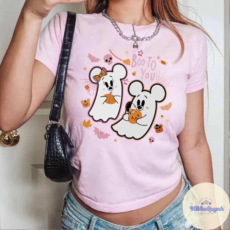May include: A light pink t-shirt featuring a Halloween-themed graphic. The graphic includes two ghost-like figures resembling cartoon mice, each holding a pumpkin. The phrase "Boo To You" is written above the figures, with bats and spiderwebs.