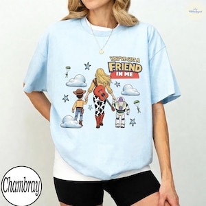 Op de afbeelding: Een lichtblauw t-shirt met een grafische illustratie van een blond personage dat hand in hand loopt met een cowboy en een space ranger. Het ontwerp bevat een gitaar, wolken, sterren, parachutespringende soldaten en de tekst "YOU'VE GOT A FRIEND IN ME."