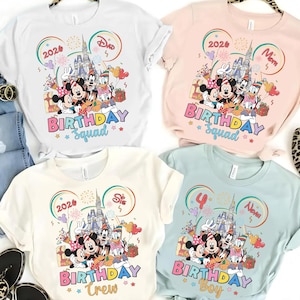 May include: Four t-shirts in white, pink, cream, and light blue, each featuring a colorful graphic of Disney characters in front of a castle. The shirts have the words "BIRTHDAY Squad", "BIRTHDAY Crew", and "BIRTHDAY Boy" printed on them.