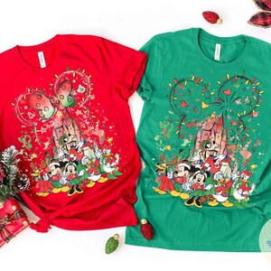 May include: Two short-sleeved t-shirts, one red and one green, featuring a festive Disney-themed design. The design includes Mickey Mouse ears decorated with Christmas lights, a castle, and various Disney characters in holiday attire.