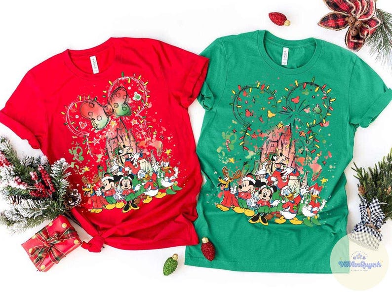 May include: Two short-sleeved t-shirts, one red and one green, featuring a festive Disney-themed design. The design includes Mickey Mouse ears decorated with Christmas lights, a castle, and various Disney characters in holiday attire.