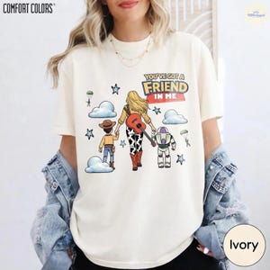 Op de afbeelding: Een ivoorkleurig t-shirt met een afbeelding van drie figuren die weglopen, waaronder een cowboy, een blond persoon met een gitaar en een space ranger. De tekst luidt "YOU'VE GOT A FRIEND IN ME" met blauwe sterren en witte wolken.