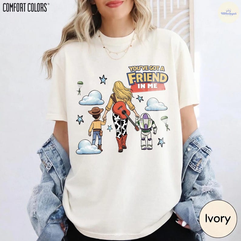 Op de afbeelding: Een ivoorkleurig t-shirt met een afbeelding van drie figuren die weglopen, waaronder een cowboy, een blond persoon met een gitaar en een space ranger. De tekst luidt "YOU'VE GOT A FRIEND IN ME" met blauwe sterren en witte wolken.