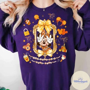 May include: A purple sweatshirt featuring a Minnie Mouse graphic in a gold frame, surrounded by festive icons like fireworks, balloons, and a castle. The design includes the text "Disneyland Park" in a decorative font.