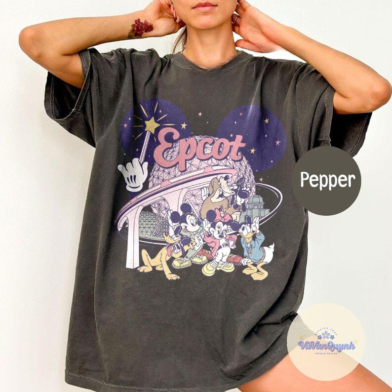 May include: Dark gray t-shirt featuring a colorful graphic of Disney characters and the word "Epcot". The design includes Mickey Mouse, Donald Duck, and Goofy, with a futuristic structure and starry background.
