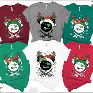 May include: A collection of Christmas-themed t-shirts in various colors, including red, green, gray, and white. Each shirt features a festive design with a Mickey Mouse head wearing a Santa hat and reindeer antlers, with the word "Name" on the design.