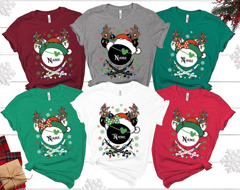 May include: A collection of Christmas-themed t-shirts in various colors, including red, green, gray, and white. Each shirt features a festive design with a Mickey Mouse head wearing a Santa hat and reindeer antlers, with the word "Name" on the design.