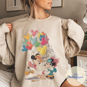 May include: Beige sweatshirt featuring a colorful graphic of Mickey and Minnie Mouse. Mickey holds a bunch of Mickey Mouse-shaped balloons. Minnie Mouse is holding a candy cane. A castle is in the background.