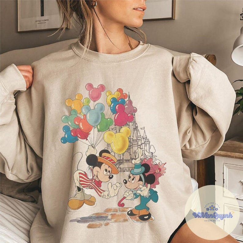May include: Beige sweatshirt featuring a colorful graphic of Mickey and Minnie Mouse. Mickey holds a bunch of Mickey Mouse-shaped balloons. Minnie Mouse is holding a candy cane. A castle is in the background.