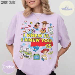 Op de afbeelding: Een lichtpaars T-shirt met korte mouwen en een grafische print van diverse Toy Story-personages, waaronder Woody, Buzz Lightyear en Mr. Potato Head. De tekst "I KNEW IT, I KNEW YOU TS" staat in geel en rood in het midden van het shirt.