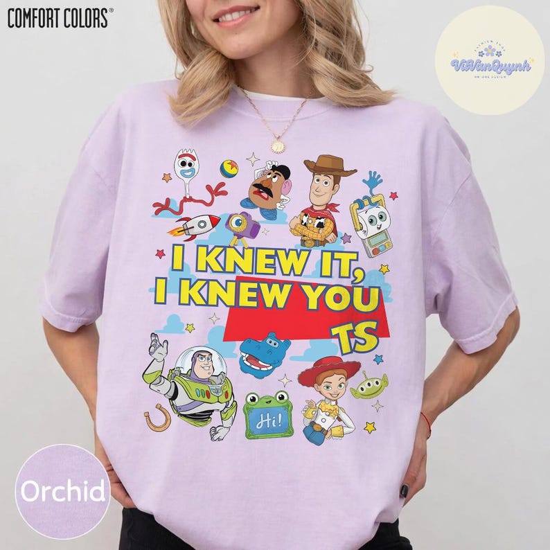 Op de afbeelding: Een lichtpaars T-shirt met korte mouwen en een grafische print van diverse Toy Story-personages, waaronder Woody, Buzz Lightyear en Mr. Potato Head. De tekst "I KNEW IT, I KNEW YOU TS" staat in geel en rood in het midden van het shirt.