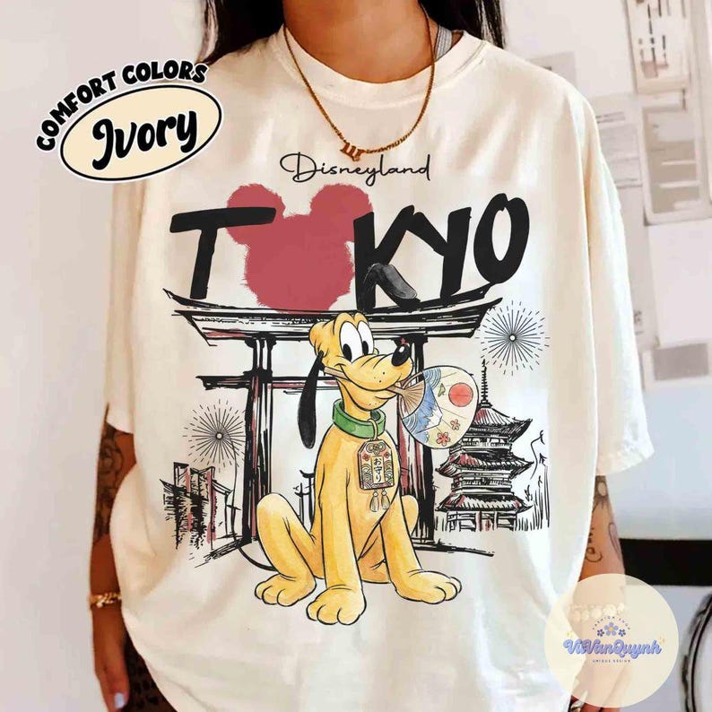 K&ouml;nnte beinhalten: Elfenbeinfarbenes T-Shirt mit einem Disneyland Tokyo-Motiv. Das Design zeigt Pluto mit einem F&auml;cher, einen roten Mickey Mouse-Kopf und japanische architektonische Elemente. Die Worte "Comfort Colors" und "Ivory" sind ebenfalls auf dem T-Shirt aufgedruckt.