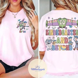 May include: Light pink t-shirt featuring a colorful graphic of characters and text from the movie Toy Story. The front of the shirt has a small graphic with the text "UF '26". The back of the shirt has the text "TO KINDERGARTEN AND BEYOND".