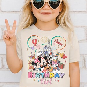 May include: A beige t-shirt featuring a colorful graphic of Disney characters, including Mickey and Minnie Mouse, Goofy, and Donald Duck. The shirt has the text "BIRTHDAY Girl" and the number "4". The design includes a castle, balloons, and confetti.