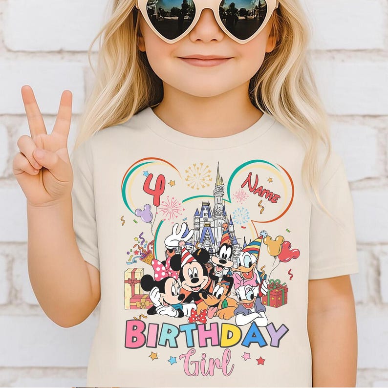 May include: A beige t-shirt featuring a colorful graphic of Disney characters, including Mickey and Minnie Mouse, Goofy, and Donald Duck. The shirt has the text "BIRTHDAY Girl" and the number "4". The design includes a castle, balloons, and confetti.