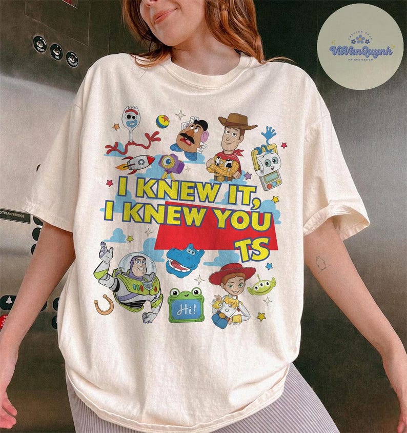 Op de afbeelding: Een cr&egrave;mekleurig t-shirt met korte mouwen en een kleurrijke grafische print van diverse geanimeerde personages uit de Toy Story-franchise. De tekst luidt I KNEW IT, I KNEW YOU TS in dikke gele en witte letters op een rode achtergrond.