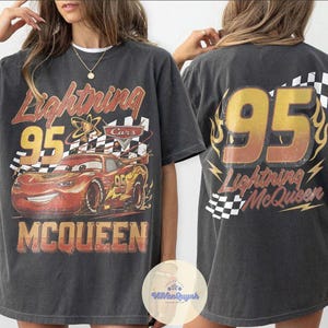 K&ouml;nnte beinhalten: Dunkelgraues T-Shirt mit einem Used-Look-Grafik von Lightning McQueen aus dem Film Cars. Auf der Vorderseite sind die Nummer 95, der Name der Figur und eine Zielflagge abgebildet. Auf der R&uuml;ckseite stehen ebenfalls die Nummer 95 und Lightning McQueens Name.