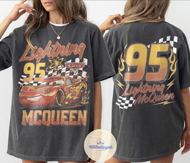 K&ouml;nnte beinhalten: Dunkelgraues T-Shirt mit einem Used-Look-Grafik von Lightning McQueen aus dem Film Cars. Auf der Vorderseite sind die Nummer 95, der Name der Figur und eine Zielflagge abgebildet. Auf der R&uuml;ckseite stehen ebenfalls die Nummer 95 und Lightning McQueens Name.