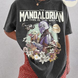 K&ouml;nnte beinhalten: Dunkelgraues T-Shirt mit einer Grafik von The Mandalorian und Grogu, umgeben von Blumen. Der Text "THE MANDALORIAN - THIS IS THE WAY" ist oben aufgedruckt. Das Shirt ist ein Comfort Colors Pepper.