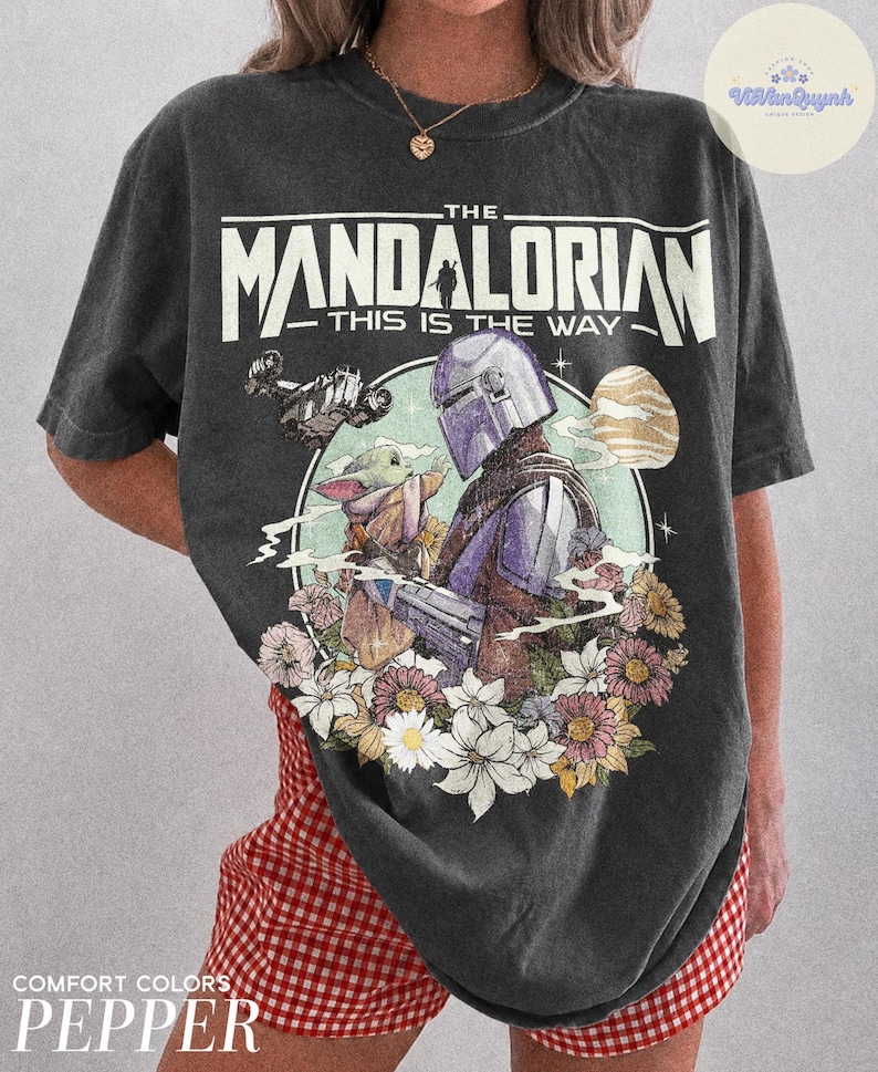 K&ouml;nnte beinhalten: Dunkelgraues T-Shirt mit einer Grafik von The Mandalorian und Grogu, umgeben von Blumen. Der Text "THE MANDALORIAN - THIS IS THE WAY" ist oben aufgedruckt. Das Shirt ist ein Comfort Colors Pepper.