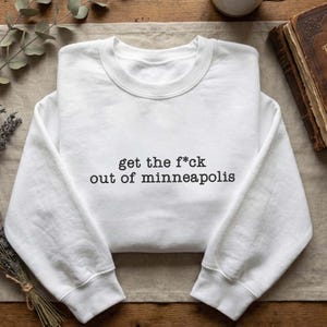 Może przedstawiać: Biała bluza z okrągłym dekoltem z czarnym napisem "get the f*ck out of minneapolis". Bluza wykonana jest z miękkiego materiału i ma długie rękawy.