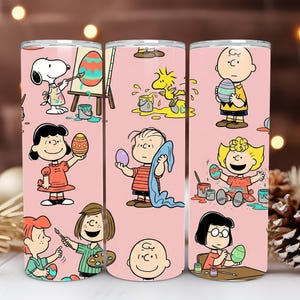 May include: Three stainless steel tumblers with a pink background featuring cartoon characters from the Peanuts gang decorating Easter eggs. Snoopy paints an egg, while Charlie Brown and Lucy hold decorated eggs.