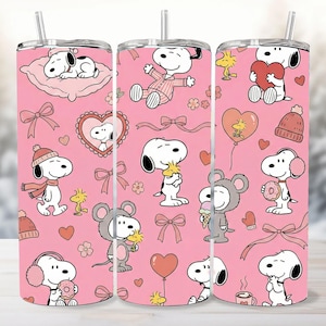 Könnte beinhalten: Drei rosa isolierte Becher mit einem sich wiederholenden Muster von Snoopy-Charakteren in verschiedenen Posen, Herzen, Schleifen und Blumen. Jeder Becher hat einen silbernen Strohhalm und einen weißen Deckel. Der Hintergrund ist eine verschwommene Winterszene.