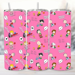 Könnte beinhalten: Drei rosa Becher mit einem sich wiederholenden Muster von Peanuts-Charakteren, Sprechblasen und dem Text "Snoopy Drops" und "Ha Ha Ha!". Jeder Becher hat einen silbernen Strohhalm.
