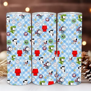 Könnte beinhalten: Drei Edelstahlbecher mit einem sich wiederholenden Muster von Snoopy-Charakteren in verschiedenen Outfits, darunter ein Astronaut, ein Dinosaurier und ein Pilot, vor einem blauen Karohintergrund. Das Design umfasst Sterne und Snoopy's Hundehütte.