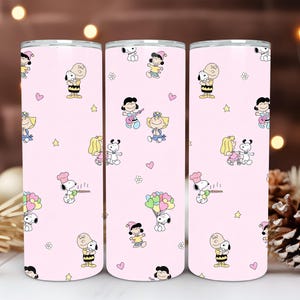 Könnte beinhalten: Drei rosa isolierte Becher mit einem sich wiederholenden Muster von Snoopy, Charlie Brown und anderen Peanuts-Charakteren. Das Design umfasst Herzen, Sterne und Blumen. Die Becher sind zylindrisch und haben einen silbernen Rand.