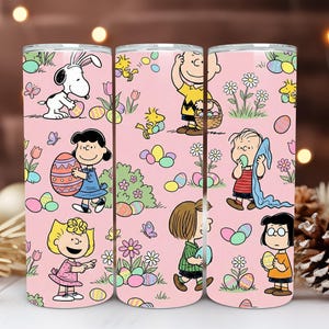 Könnte beinhalten: Drei rosa isolierte Becher mit Peanuts-Charakteren und Ostereiern. Snoopy mit Hasenohren, Charlie Brown mit einem Korb und Lucy mit einem Osterei sind unter den Figuren. Das Design umfasst Blumen, Schmetterlinge und bunte Eier.