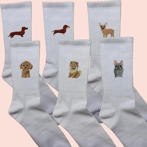 Könnte beinhalten: Weiße Sportsocken mit gestickten Hunde-Designs. Die Socken zeigen verschiedene Hunderassen, darunter Dackel, Chihuahua, Pudel, Chow Chow und Französische Bulldogge. Die Hunde-Stickerei ist in Braun-, Beige- und Grautönen gehalten.