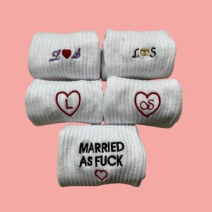 Könnte beinhalten: Weiße Armbänder mit gestickten Designs. Einige Armbänder zeigen Initialen mit Herzen, während andere den Text "MARRIED AS FUCK" mit einem Herzen anzeigen. Die Armbänder sind vor einem rosa Hintergrund gestapelt.