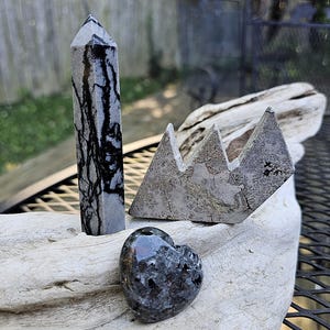 May include: A collection of polished stones, including a tall, gray obelisk with black veining, a heart-shaped stone, and a triangular mountain-shaped stone. The stones are displayed on a piece of driftwood.